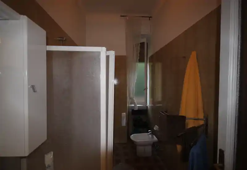 Bagno con cabina doccia, bidet, portasciugamani e una piccola finestra; piastrelle marroni e luce naturale moderata.
