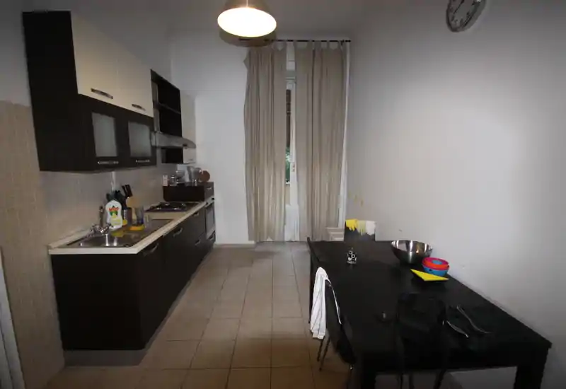 Cucina a corridoio moderna con mobili inferiori scuri e pensili chiari, forno incassato e piano cottura a gas; piccolo tavolo da pranzo lungo la parete e finestra con tende che fornisce luce naturale.