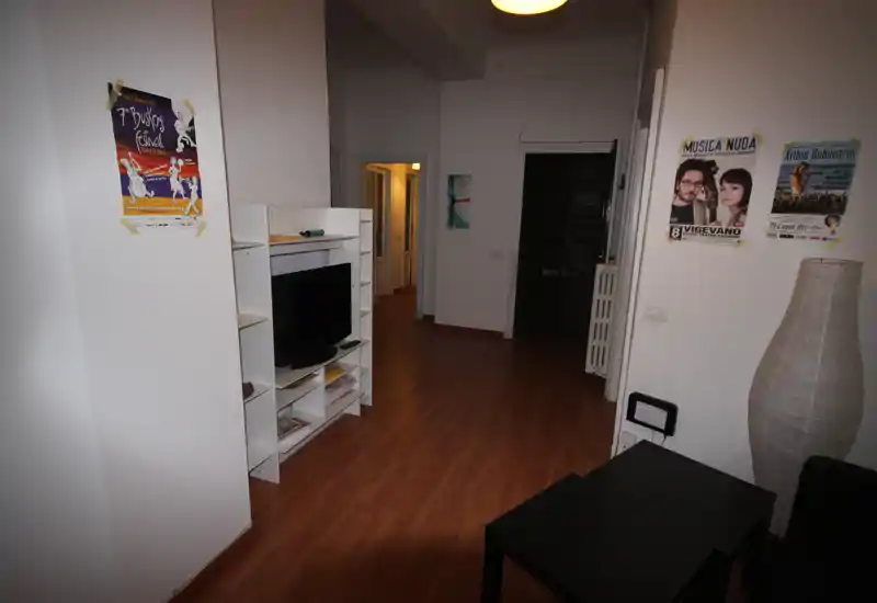 Area giorno semplice e luminosa con mobile TV, mensole e pavimento effetto legno; si vede il corridoio e l'ingresso sullo sfondo.
