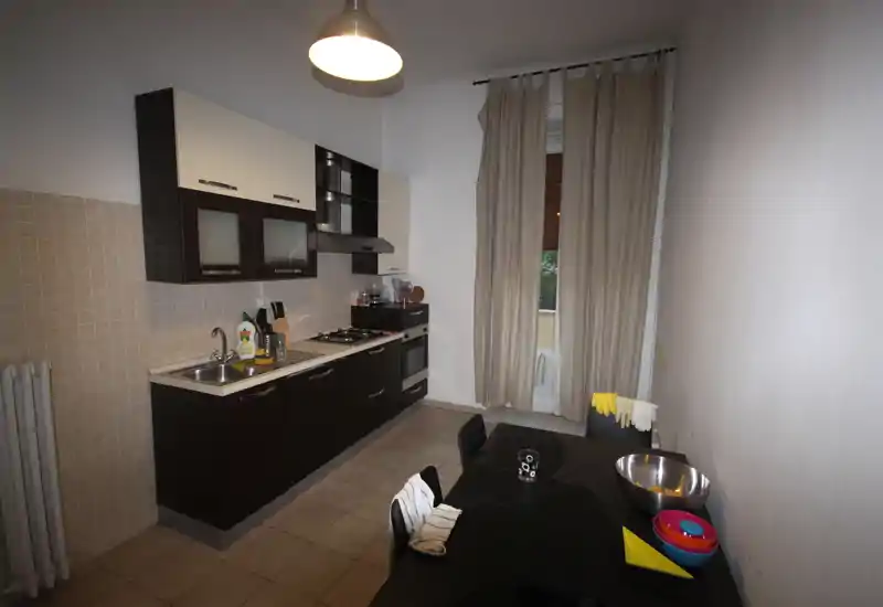 Cucina moderna a corridoio con basi scure e piano chiaro, forno incassato e piano cottura a gas, lavello in acciaio e piccolo tavolo da pranzo vicino a una finestra con tenda. Layout pulito e funzionale per l'uso quotidiano.
