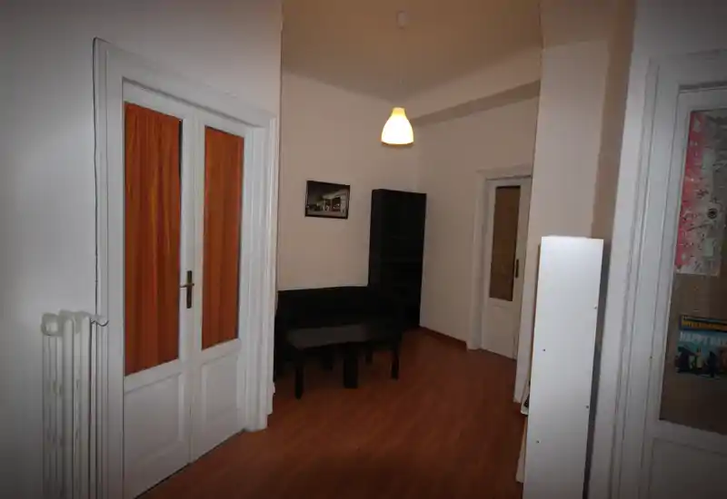Soggiorno minimale con un piccolo divano, tavolino e scaffale; pavimento in legno e lampada a sospensione creano uno spazio salotto compatto e funzionale.