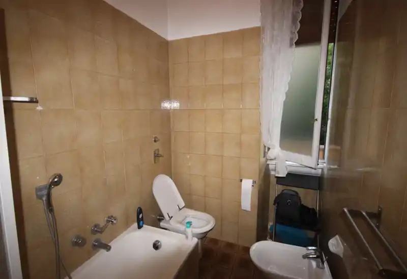 Bagno compatto con vasca, bidet e WC; pareti con piastrelle beige e finestra con tenda in pizzo che fornisce luce naturale.