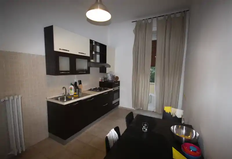 Cucina moderna a corridoio con mobili bassi scuri e pensili chiari, forno e piano cottura a gas integrati, tavolo da pranzo in primo piano e una finestra alta con tende che fornisce luce naturale.