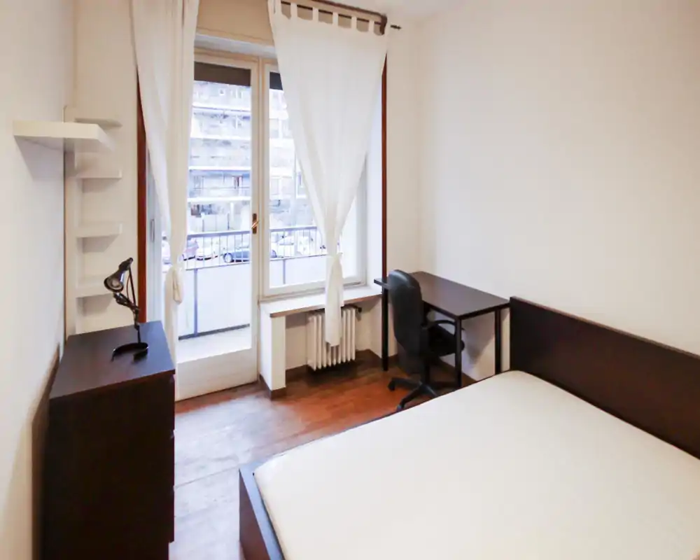 Dormitorio luminoso y sencillo con cama doble, escritorio y gran ventana con acceso a un pequeño balcón. Decoración neutra y suelo de madera que aportan un ambiente ordenado y funcional.