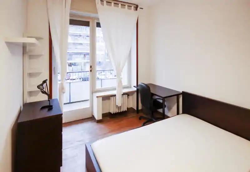 Dormitorio luminoso y sencillo con cama doble, escritorio y gran ventana con acceso a un pequeño balcón. Decoración neutra y suelo de madera que aportan un ambiente ordenado y funcional.