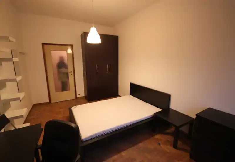 Dormitorio ordenado con cama doble, mesitas, armario y escritorio. Iluminación cálida desde arriba y suelo de madera crean un espacio simple y funcional, ideal para anuncios de alquiler.