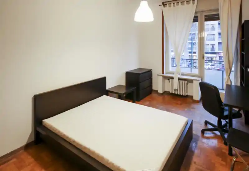 Dormitorio sencillo y luminoso con cama doble, mesita y cómoda. Ventana grande con cortinas que aporta luz natural y una pequeña zona de trabajo con silla.