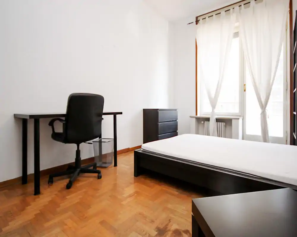 Dormitorio luminoso y minimalista con cama baja, muebles negros sencillos y un escritorio junto a la pared. Gran ventana con cortinas translúcidas que aporta mucha luz natural.