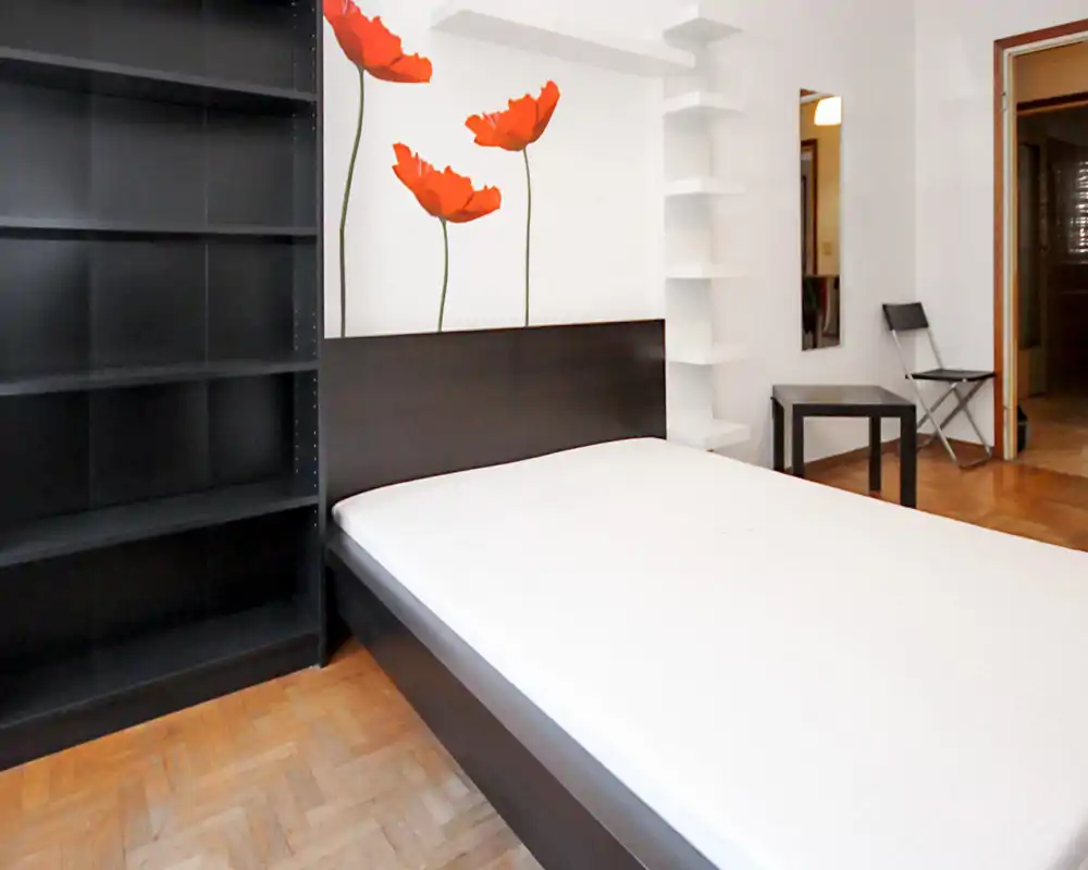 Dormitorio moderno y luminoso con cama doble grande, cabecero oscuro y estanterías minimalistas; vinilos de amapolas en la pared y suelos de parquet.
