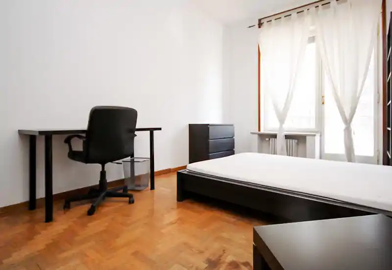 Bright, minimalist bedroom with a low platform bed, simple black furnishings and a work desk by the wall. Large window with sheer curtains provides abundant natural light.