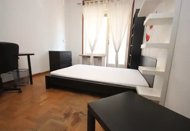 Bright, simply furnished bedroom with a low black bed frame, mattress and natural light from a large window with sheer curtains. Minimalist furniture includes a desk, drawers and wall shelving on parquet flooring.