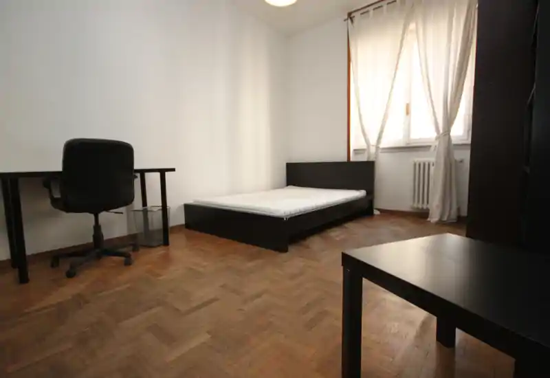 Dormitorio sencillo y bien iluminado con una cama baja negra y colchón, escritorio junto a la pared; suelo de parquet y ventana con cortinas que aportan luz natural cálida.