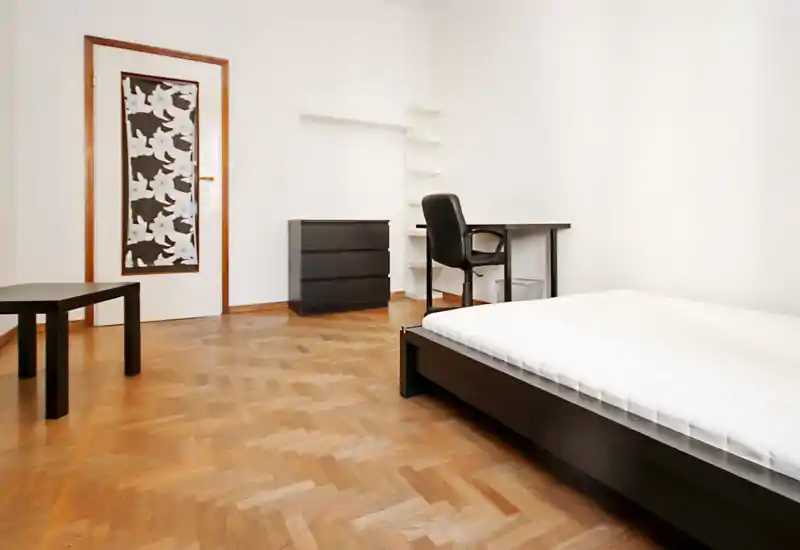 Dormitorio minimalista con estructura de cama baja negra y colchón blanco, escritorio con silla, cómoda y suelo de parquet; luminoso y amueblado de forma sencilla, ideal para una persona.