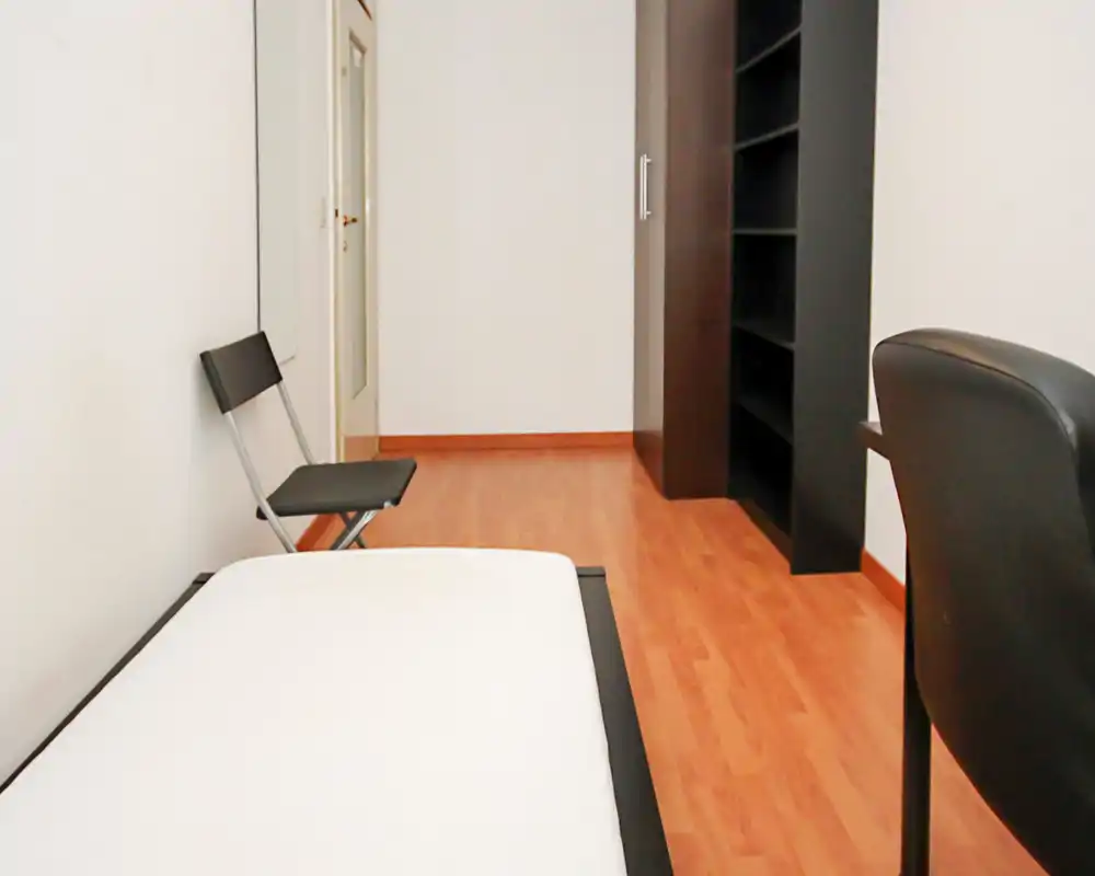 Camera da letto minima con letto singolo visibile in primo piano, una sedia pieghevole, uno specchio a parete e una libreria alta scura; pavimento in laminato pulito e pareti bianche neutre creano un ambiente semplice e ordinato.