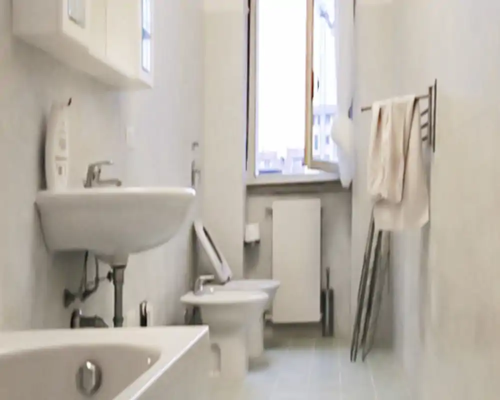 Baño compacto y limpio con bañera, lavabo mural, bidé y WC; la luz natural suave desde la ventana genera un ambiente luminoso y neutro.