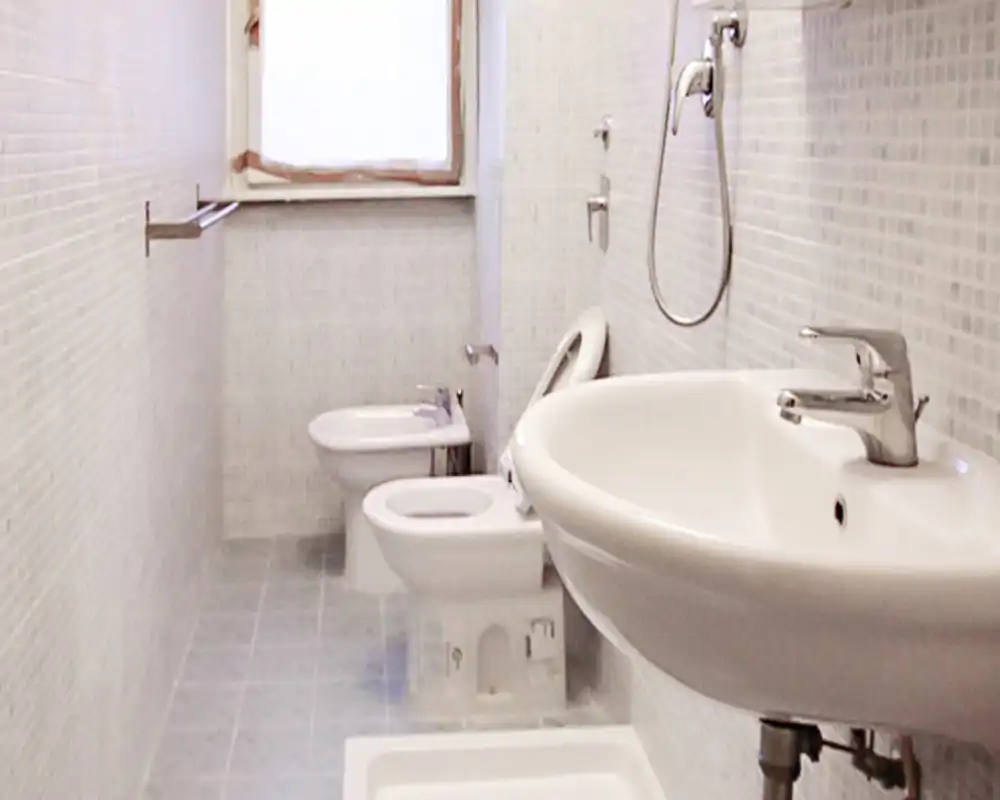 Baño limpio y estrecho con lavabo, inodoro y bidé visibles; azulejos de mosaico claros y una ventana que aporta luz natural.