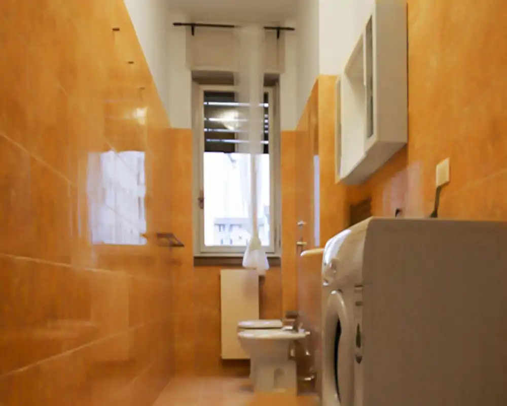 Baño pequeño con azulejos naranja, inodoro y lavadora, iluminado suavemente por la luz natural de la ventana.