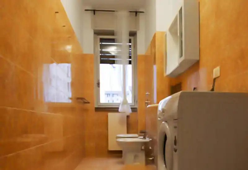 Small bathroom with warm orange tiles, a toilet and a washing machine under soft natural light from a window.
