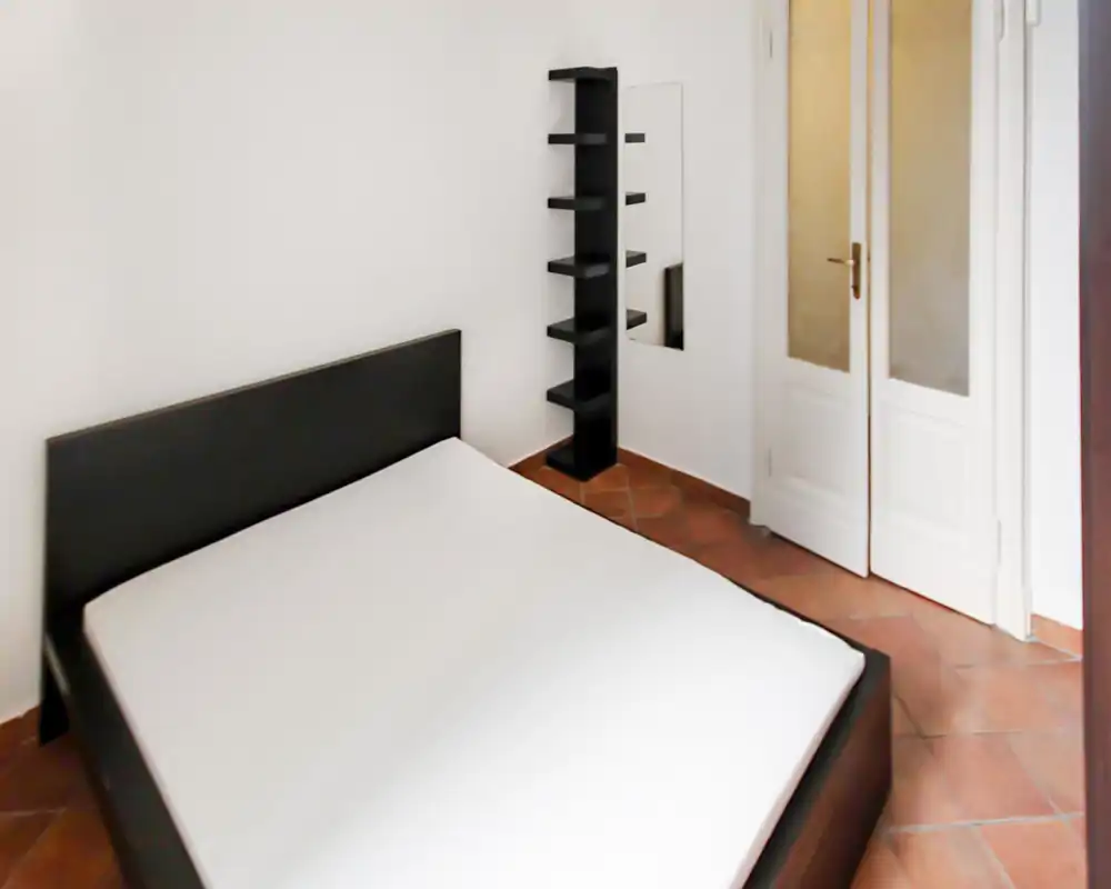 Dormitorio minimalista y luminoso con cabecero oscuro y colchón sobre estructura baja, suelo de barro cocido y estantería alta junto a un espejo.