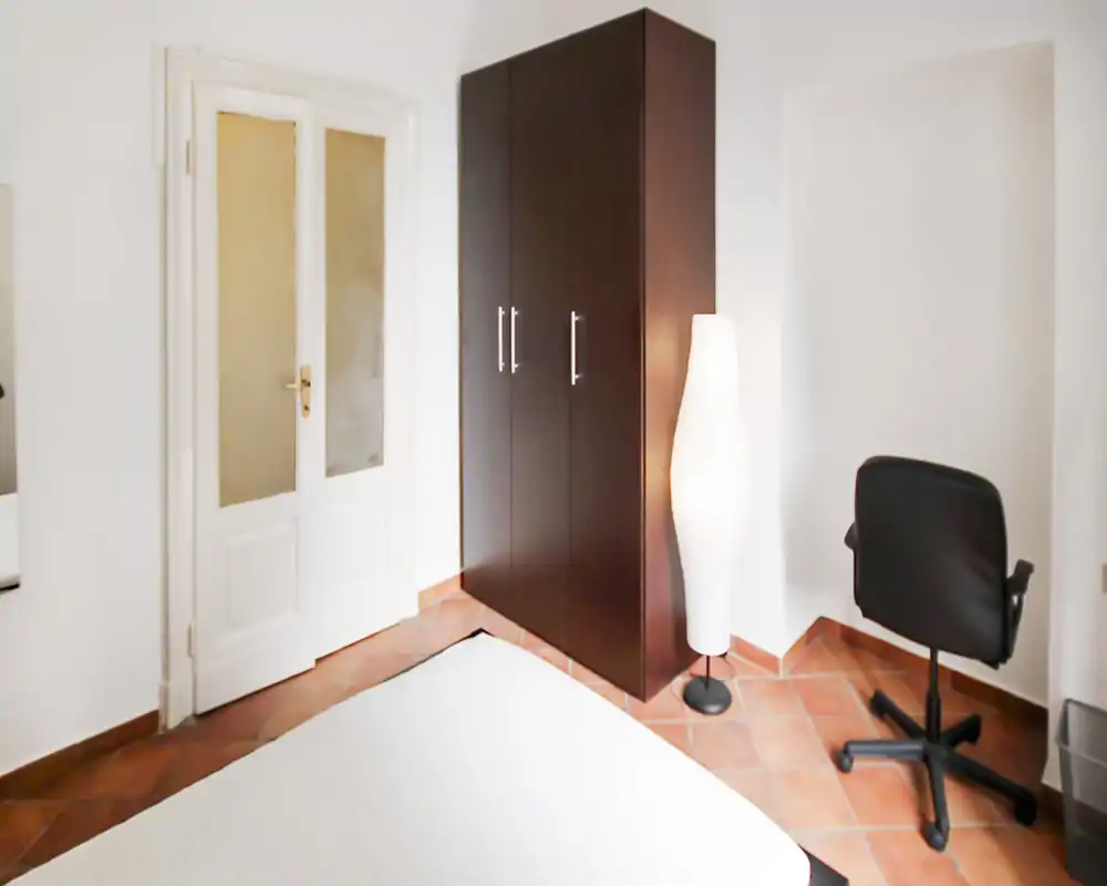 Rincón de dormitorio luminoso y sencillo con colchón en primer plano, armario oscuro, lámpara de pie y silla giratoria sobre suelo de terracota.