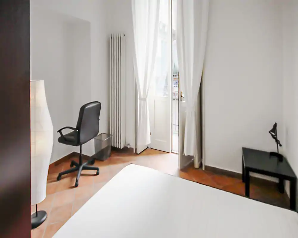 Dormitorio luminoso y minimalista con colchón visible en primer plano, escritorio y silla junto a la ventana y cortinas translúcidas que dan a un pequeño balcón.