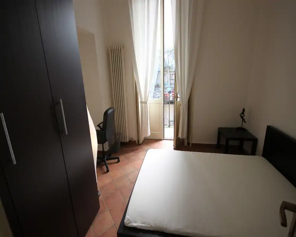 Dormitorio luminoso y sencillo con cama doble, mesita y armario; la puerta al balcón proporciona luz natural agradable.