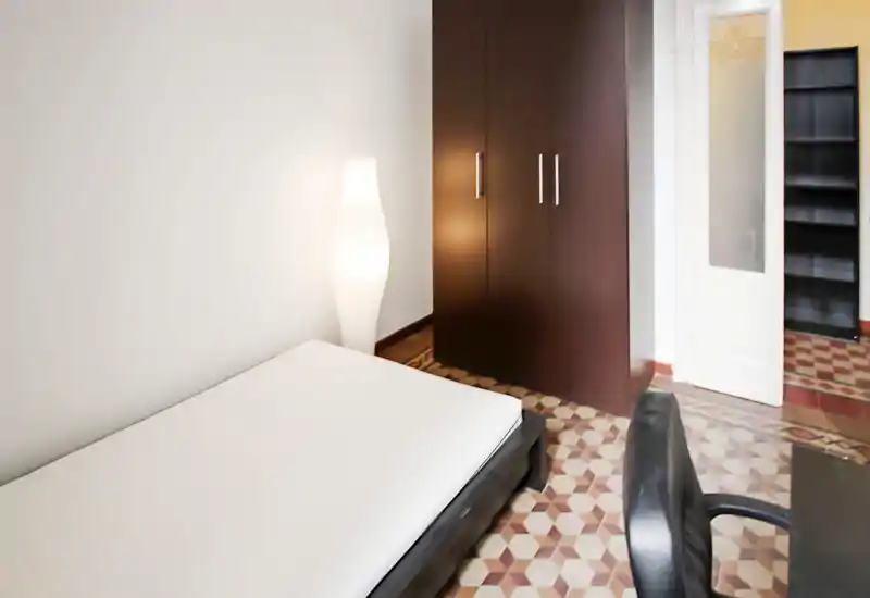 Camera da letto pulita e arredata in modo essenziale con materasso su rete bassa, lampada da terra e armadio in legno scuro. Pavimento a piastrelle decorate e pareti neutre creano un ambiente luminoso e tranquillo.