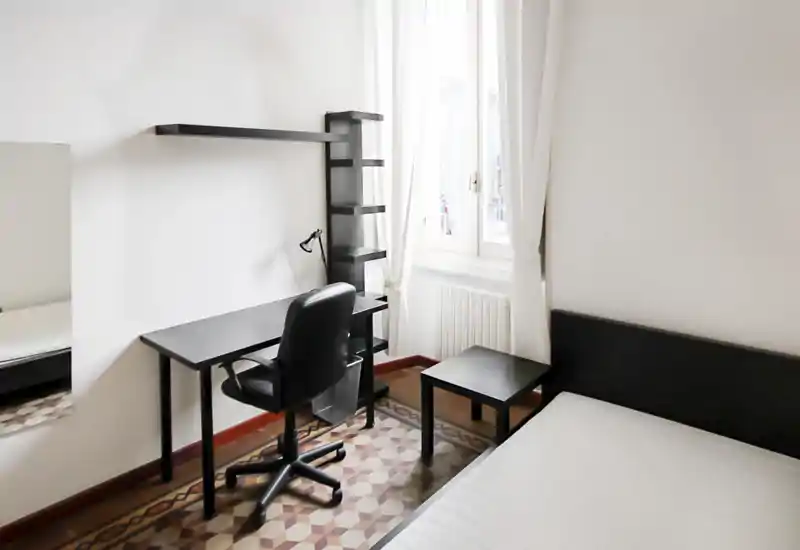 Camera da letto luminosa e minimal con letto singolo, testiera nera e una scrivania compatta vicino alla finestra. Linee pulite, piastrelle decorative e luce naturale rendono l’ambiente arioso e funzionale.