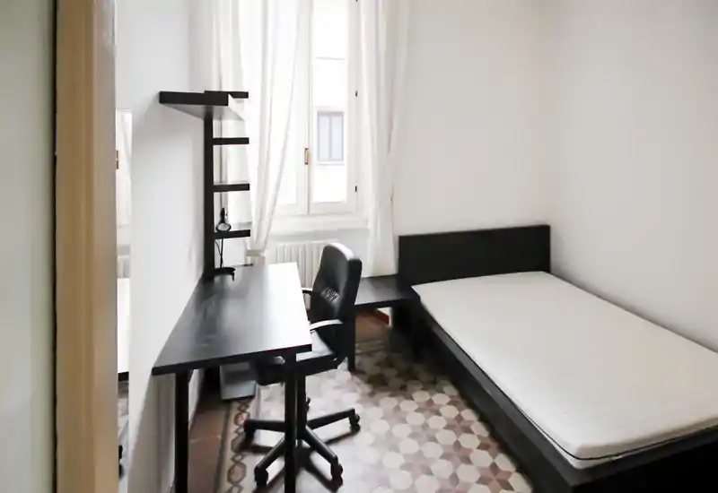 Camera singola minimal e luminosa con letto basso nero, scrivania e sedia coordinati, luce naturale da una finestra ampia e pavimento piastrellato decorato — spazio funzionale ideale per studenti o soggiorni brevi.