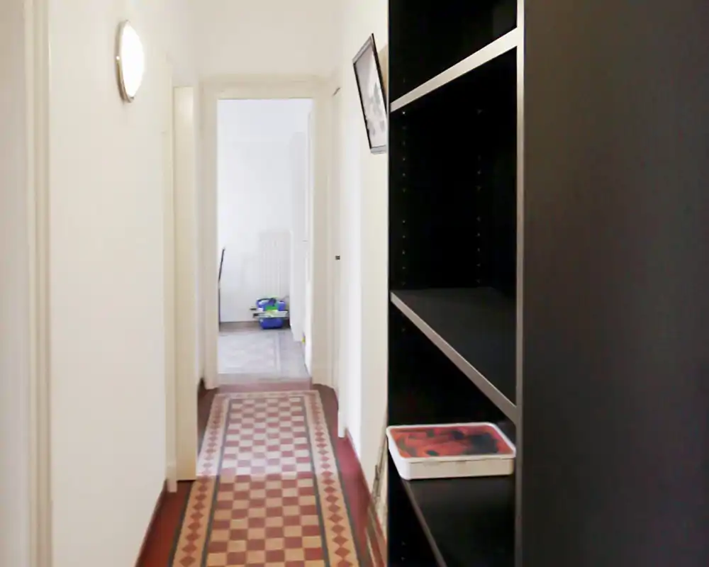 Corridoio stretto e ben illuminato con pavimento a piastrelle decorative e una libreria sul lato destro — vista chiara dell'ingresso/corridoio che conduce alle stanze adiacenti.