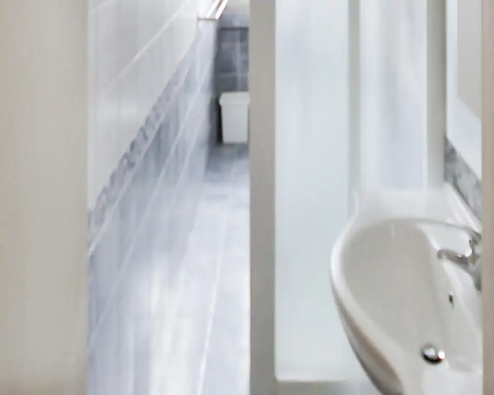 Bagno stretto a corridoio con un lavabo bianco in primo piano e pavimento e rivestimenti in piastrelle lungo un layout lungo e sottile.