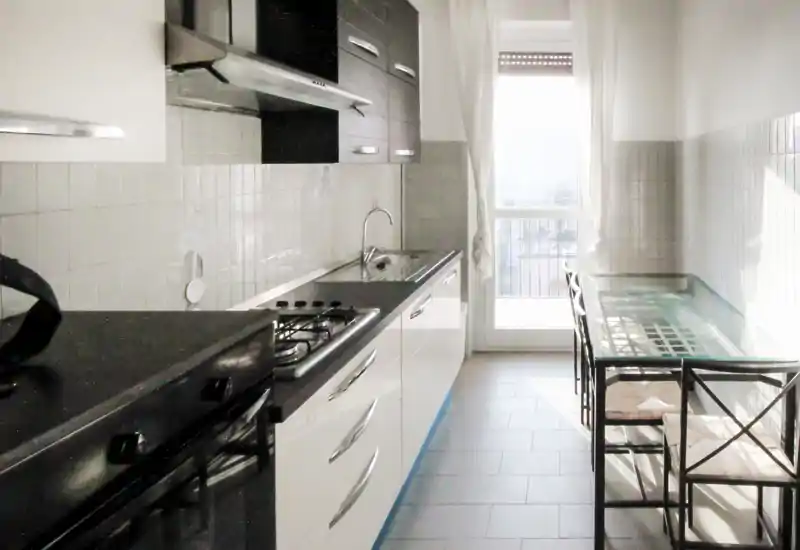 Cucina a corridoio luminosa con mobili moderni, piano cottura a gas, lavello e un tavolo in vetro vicino alla porta del balcone che lascia entrare luce naturale.
