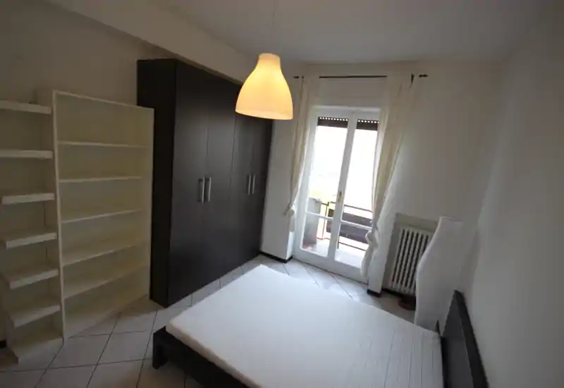 Chambre moderne et lumineuse avec lit double, armoire et étagères ; portes donnant sur le balcon apportant une belle lumière naturelle et une atmosphère épurée.