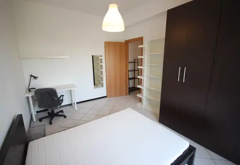 Chambre lumineuse et simplement meublée avec lit double, grand placard foncé, étagères ouvertes et petit bureau contre le mur — agencement soigné et fonctionnel.