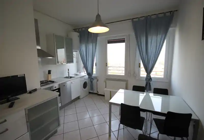 Bright, compact kitchen with integrated appliances, a dining table for four and large windows with blue curtains providing natural light.
