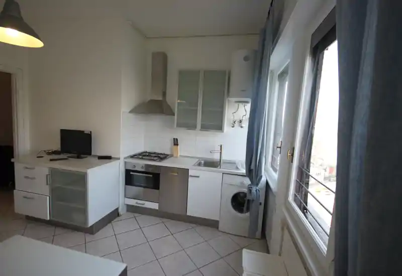 Kitchenette moderna y compacta con horno integrado, placa de gas, fregadero y lavadora bajo encimera; grandes ventanas con cortinas que aportan buena luz natural.