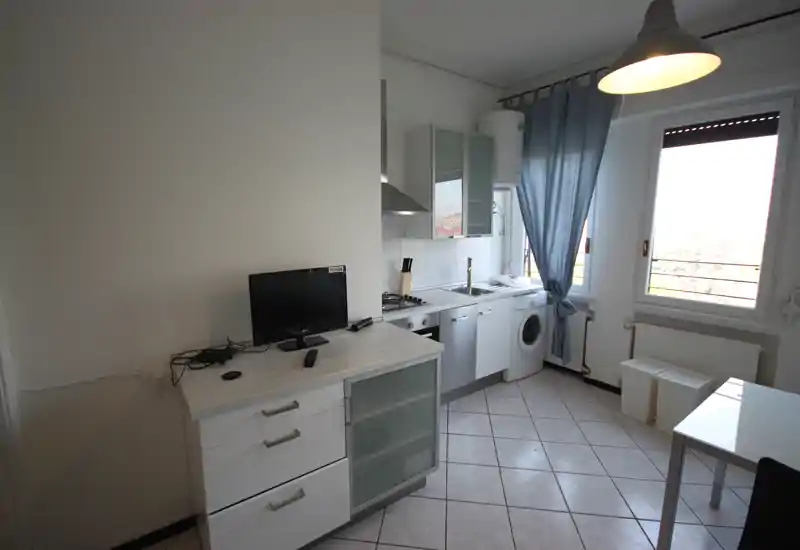 Compact, modern kitchen with white cabinetry, built-in stove, sink and washing machine next to a bright window with blue curtains.