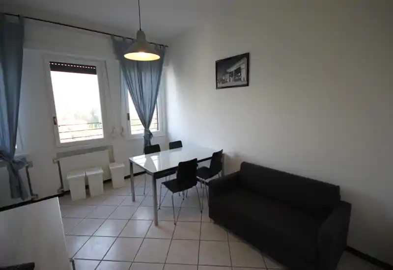 Small, modern living area with a compact dark sofa, glass-top dining table and four chairs placed by two large windows that provide plenty of natural light.