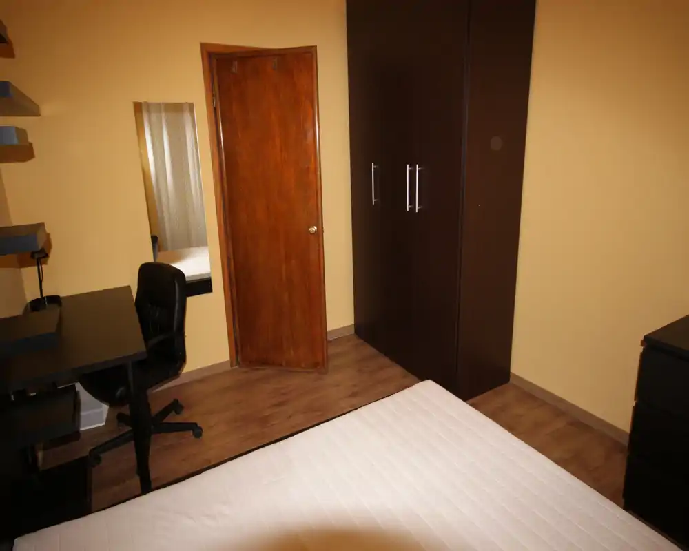 Camera da letto semplice con letto matrimoniale visibile, armadio e scrivania. L’illuminazione calda e il pavimento in legno donano un’atmosfera accogliente e pratica.