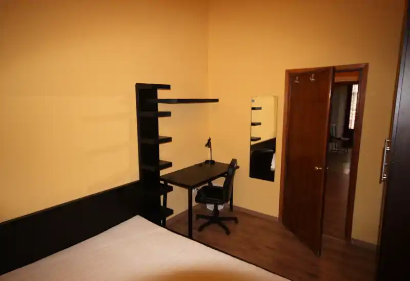 Camera da letto semplice con angolo del letto visibile, testiera scura e scrivania con mensole nell'angolo. Pareti gialle calde e pavimento in legno creano un ambiente accogliente e ben illuminato, adatto a un annuncio di affitto.