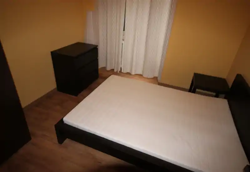 Camera da letto semplice e ordinata con letto matrimoniale su struttura in legno scuro, comodino e cassettiera coordinati e tende chiare per la privacy.