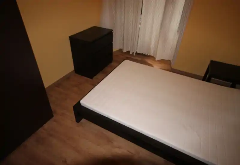 Camera da letto semplice e ordinata con un letto matrimoniale su struttura scura ben visibile, pavimento in laminato e mobili abbinati; tende leggere filtrano la luce.
