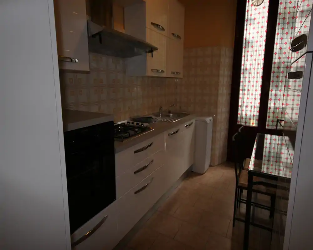 Cucina compatta con pensili bianchi, piano cottura a gas, forno e lavello; porta con vetro decorato che lascia entrare luce naturale. Perfetta per un appartamento piccolo.