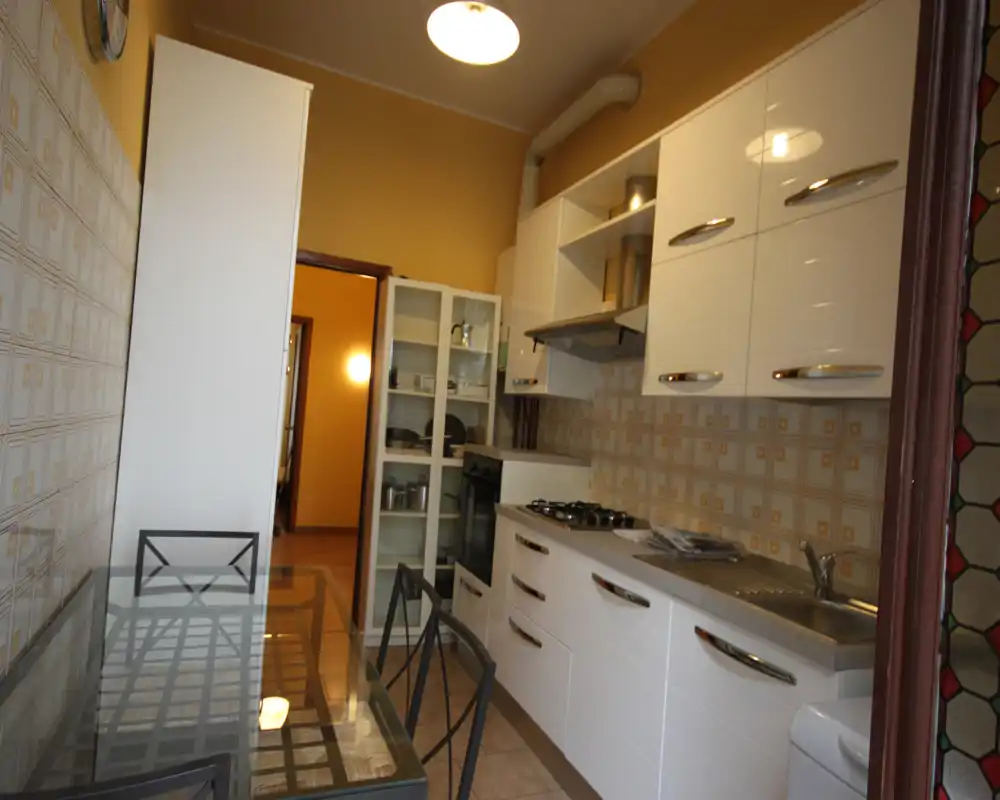 Cucina luminosa e pratica con mobili bianchi, piano cottura a gas, lavello in acciaio e tavolo in vetro con sedie in metallo — ordinata e funzionale per cucinare e pranzare.