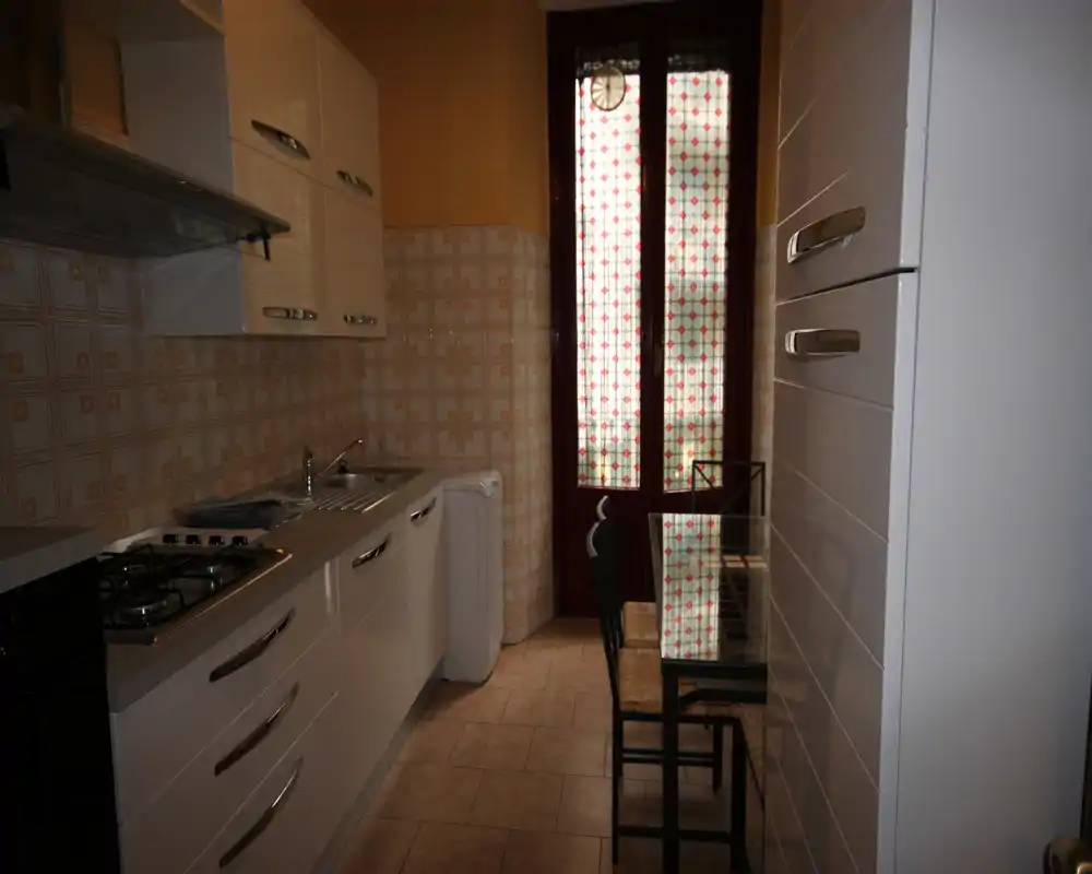 Cucina stretta con mobili bianchi moderni, piano cottura a gas, lavello e un piccolo tavolo in vetro accanto a una porta con vetro decorato che filtra la luce.