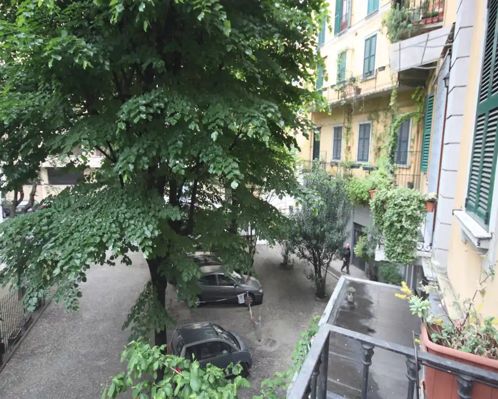 Veduta di un cortile ombreggiato con auto parcheggiate, alberi maturi e balconi di appartamenti circostanti; uno spazio esterno urbano con vegetazione.