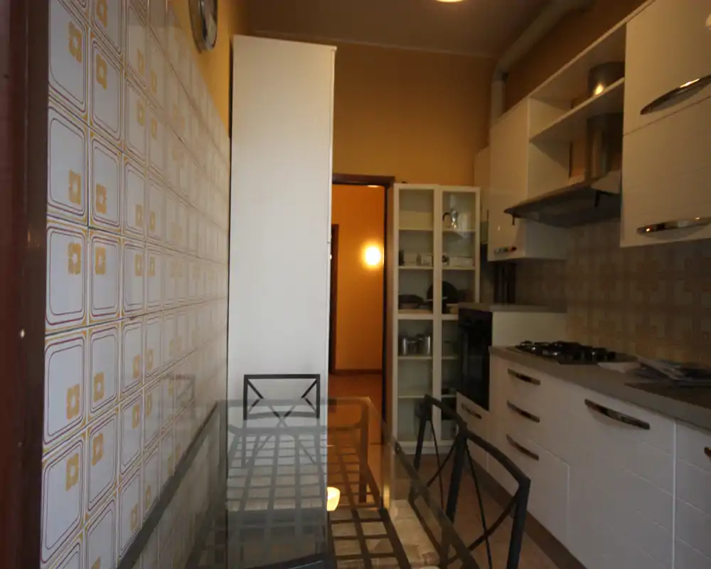 Cucina attrezzata con tavolo da pranzo in vetro, mobili bianchi moderni e pareti rivestite da piastrelle decorative; piano cottura e armadietti visibili per un ambiente funzionale.