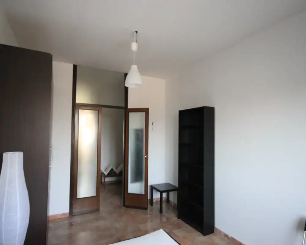 Camera da letto semplice e minimale: angolo del materasso visibile, libreria alta e tavolino. Pareti neutre e luce naturale rendono lo spazio adatto per un annuncio in affitto, con focus su metratura e contenimento.