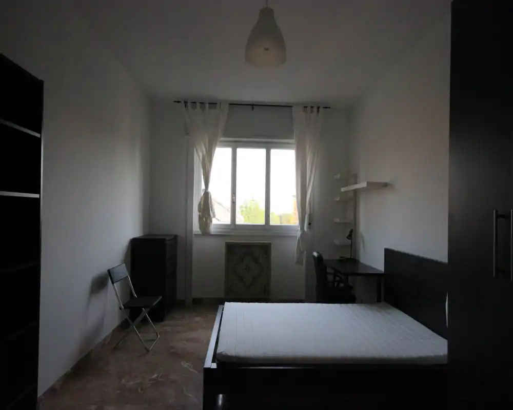 Camera da letto minimale e ordinata con letto matrimoniale, scrivania vicino alla finestra e luce naturale; tonalità neutre e arredi moderni semplici creano un'atmosfera tranquilla adatta agli annunci.