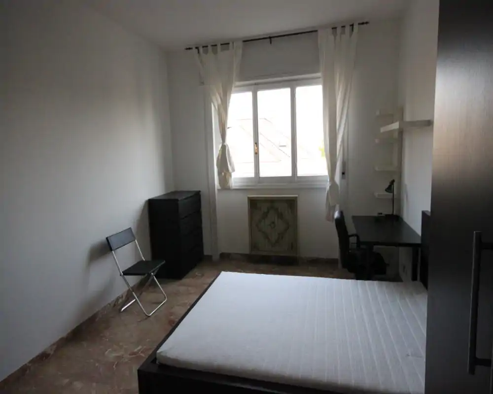 Camera da letto luminosa e arredata con semplicità: letto matrimoniale, scrivania e comò vicino a una grande finestra che illumina l'ambiente.